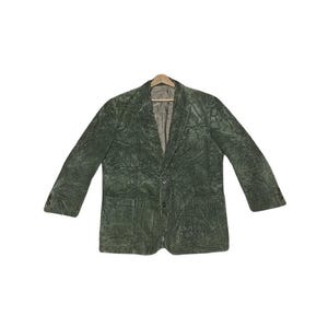 Peut inclure: Un blazer en velours côtelé vert avec une fermeture à un bouton et deux poches avant. La veste est suspendue à un cintre en bois sur fond blanc. Le blazer a un aspect texturé et des manches longues.