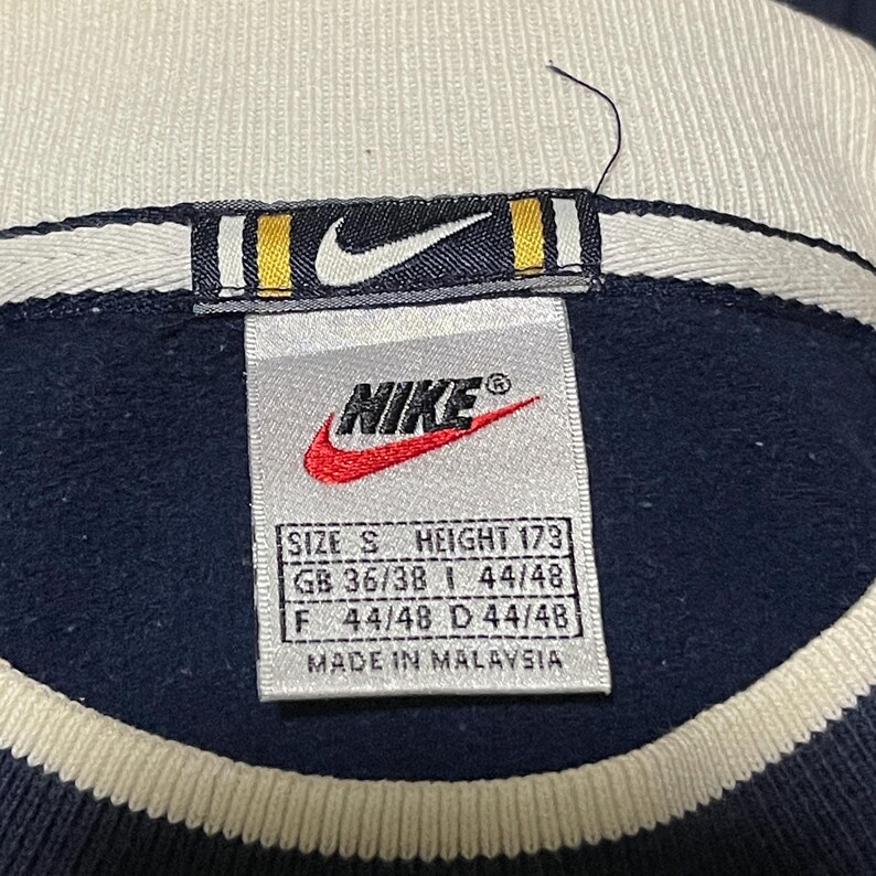 Puede incluir: Primer plano de una etiqueta de sudadera Nike azul marino. La etiqueta presenta el logotipo de Nike en blanco y rojo, con detalles de talla y medidas. La etiqueta dice "SIZE S HEIGHT 173" y "MADE IN MALAYSIA".