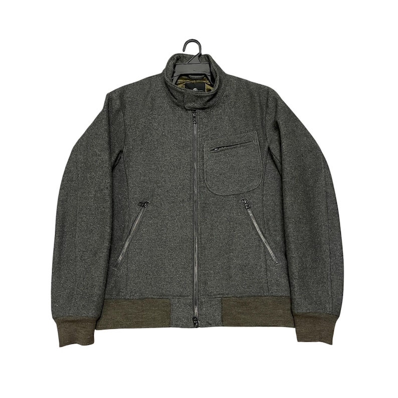 May include: A charcoal gray wool jacket with a full-length zipper, a small chest pocket, and two zippered side pockets. The jacket has a ribbed, olive green hem and cuffs, and a stand-up collar. The jacket is hanging on a black hanger.