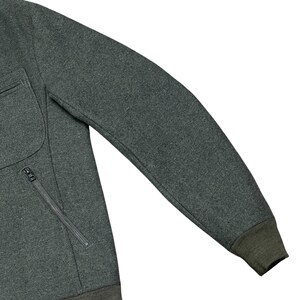 May include: Close-up of a dark gray sweatshirt with a zippered pocket and a dark green ribbed cuff. The sweatshirt has a textured appearance and a simple, classic design. The sleeve is extended, showcasing the garment's shape.