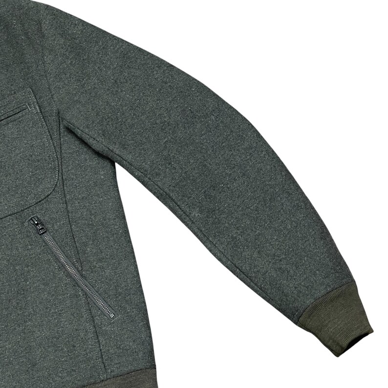 May include: Close-up of a dark gray sweatshirt with a zippered pocket and a dark green ribbed cuff. The sweatshirt has a textured appearance and a simple, classic design. The sleeve is extended, showcasing the garment's shape.