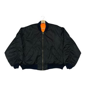 Può includere: Giacca bomber nera con chiusura a zip, due tasche frontali e colletto e polsini a coste blu navy. La giacca ha una fodera arancione ed è appesa a una gruccia di legno.