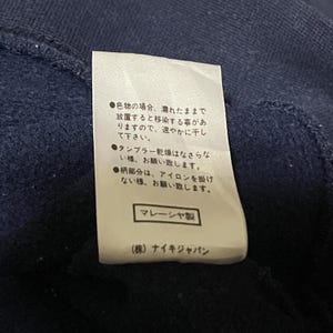 Puede incluir: Una etiqueta blanca con texto japon&eacute;s y un fondo azul marino. La etiqueta incluye instrucciones de cuidado y el texto "マレーシヤ製" y "(株)ナイキジャパン".