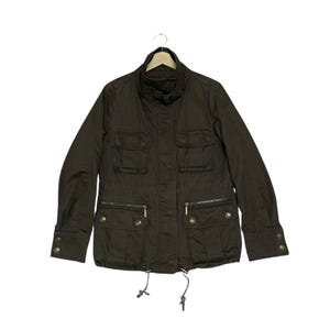 May include: A dark brown jacket with multiple pockets, a stand-up collar, and a drawstring waist. The jacket has long sleeves with buttoned cuffs and a zip closure. The jacket is hanging on a wooden hanger.