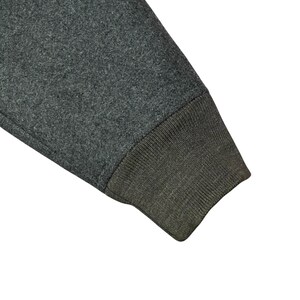 May include: Close-up of a gray sweatshirt sleeve with a ribbed, olive green cuff. The fabric has a textured appearance, and the sleeve is angled, showing the seam where the cuff is attached.