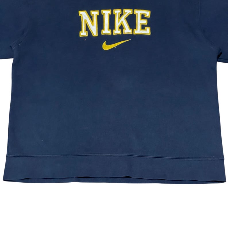 Puede incluir: Sudadera azul marino con la palabra "NIKE" en letras amarillas en el pecho. Un logotipo de Nike amarillo est&aacute; debajo. La prenda es de color s&oacute;lido.