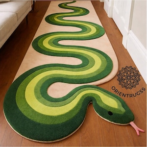 Puede incluir: Una alfombra alargada con un diseño de serpiente sinuosa en tonos verde oscuro, verde bosque y verde lima sobre un fondo beige. La serpiente tiene una cabeza definida con un ojo negro y una lengua bífida rosa. El texto ORIENTRUGSS es visible.