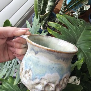May include: A handcrafted ceramic mug with a distinctive, textured pattern. The mug has a light blue and cream glaze with a brown rim. The handle is part of the design. The mug is held in front of vibrant green plants.