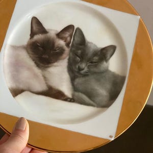 May include: A decorative plate featuring a painting of two cats. One cat is a Siamese with brown and cream fur, and the other is a gray cat. The plate has a white center with a gold rim and gold square accents.