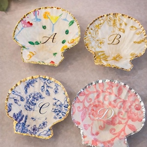 May include: Four decorative seashell-shaped plates with gold or silver trim. Each plate features a different design: a cocktail, floral patterns, and the letters A, B, C, and D. The plates are white with colorful accents.