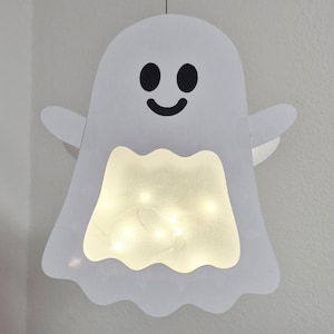 Puede incluir: Una decoración de pared con forma de fantasma blanco que presenta una cara sonriente con ojos ovalados negros y una boca curva. El centro del fantasma tiene una sección recortada iluminada por luces cálidas, creando un brillo suave.