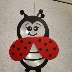 Op de afbeelding: Een hangende lieveheersbeestje decoratie met een zwarte kop, witte gezichtsdetails, grote ogen en een glimlachende mond. Het lichaam heeft twee felrode vleugels met zwarte stippen en een wit onderste gedeelte met zwarte horizontale strepen.