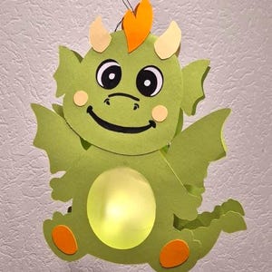Può includere: Una decorazione in carta a forma di drago verde con una pancia circolare giallo brillante, accenti arancioni sulla testa e sui piedi e corna giallo pallido. Il drago ha grandi occhi bianchi e neri, una bocca sorridente e ali laterali.
