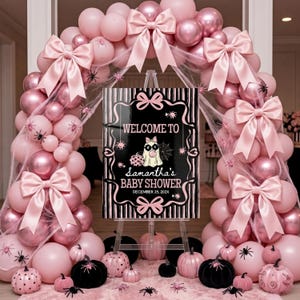 May include: A baby shower sign on an easel with a black and white striped background, pink bows, and a ghost illustration. The text reads: WELCOME TO Samantha's BABY SHOWER DECEMBER 25, 202X. The sign is framed by a large arch of pink balloons and spiders.