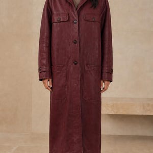 May include: A long, deep burgundy coat featuring a classic collar, a front button-down closure, two chest pockets with buttoned flaps, and two large lower patch pockets. The coat has long sleeves with buttoned cuffs and a straight, structured fit.