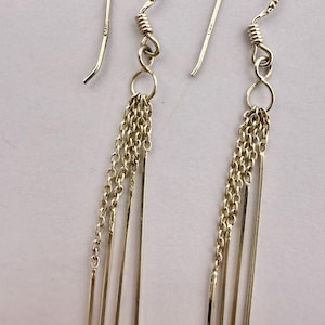 May include: A pair of silver-toned dangle earrings featuring a curved ear wire hook connected to a small circular ring. From each ring hang three delicate, thin metal chains and two slender, straight metal bars of matching silver-toned finish.