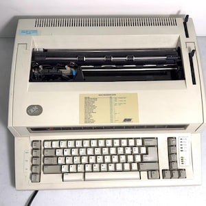 May include: A beige IBM electronic typewriter featuring a QWERTY keyboard, a black platen roller, and a central Quick Reference Guide sticker. The device includes a top-mounted paper feed slot, function keys on the left, and status indicator lights on the right side of the keyboard panel.