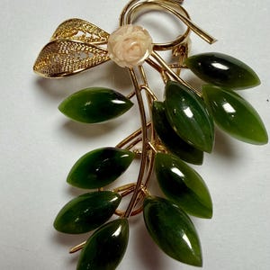 May include: A gold-toned brooch featuring a floral and leaf design. The brooch has a carved pink rose, green jade leaves, and a filigree accent. The leaves are oval-shaped and arranged along a gold stem.