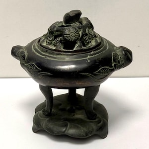 May include: A dark bronze incense burner featuring a rounded body with embossed floral motifs and two side handles. The lid is intricately pierced with a floral design and topped with a finial, all resting on three curved legs and a base.