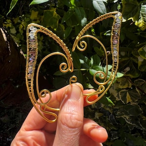 May include: A gold-toned wire ear cuff featuring intricate swirling metal patterns and a row of small, clear, faceted beads along the upper edges. The structure is shaped like pointed, wing-like forms with delicate wire wrapping and loops.