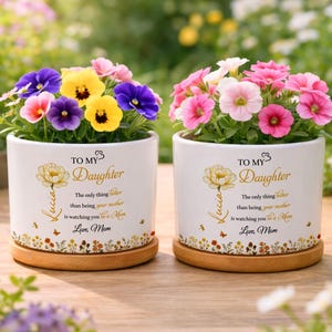 Puede incluir: Dos macetas de cerámica blanca con flores coloridas, cada una sobre un platillo de madera. Las macetas tienen el texto "To My Daughter" y un diseño floral. Las flores incluyen flores rosas, moradas y amarillas.