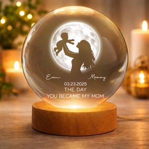 May include: A clear glass sphere with a silhouette of a mother and child against a moonlit background. The sphere sits on a wooden base and has the text "Evan", "Mommy", "03.23.2025", and "THE DAY YOU BECAME MY MOM".