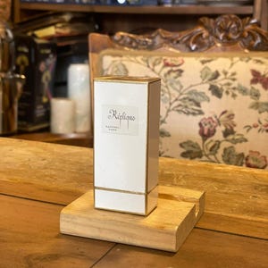 May include: A white rectangular perfume box with gold trim, labeled "Réplique" by Raphael Paris, sits on a small wooden stand. The background includes a wooden table, a floral patterned chair, and shelves with various items.