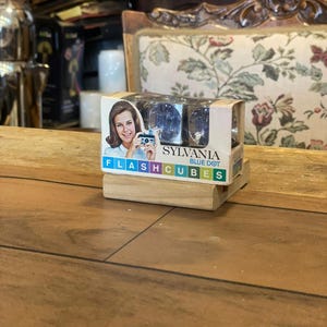 May include: A vintage Sylvania Blue Dot Flashcubes box with a woman holding a camera on the front. The box contains four blue flashcubes. The box is on a wooden block, with a wooden table in the foreground.