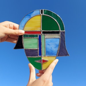 May include: A colorful stained glass art piece in the shape of a face, held against a bright blue sky. The design features geometric shapes in yellow, green, blue, red, and purple, outlined with silver solder. The piece has a unique, abstract aesthetic.