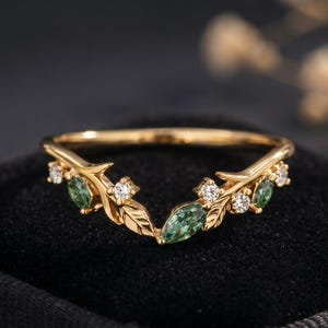 May include: A gold-colored ring with a leaf and vine design. The ring features green oval-cut gemstones and small, round, clear gemstones. The ring is set against a black background.