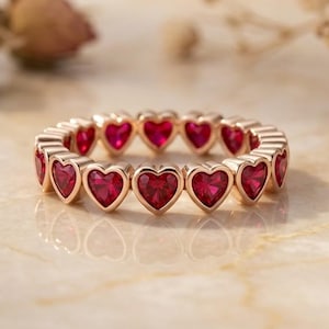 Puede incluir: Un anillo de eternidad en oro rosa que presenta una fila continua de vibrantes piedras preciosas rojas en forma de corazón. El anillo está elaborado con un acabado de metal pulido, mostrando una serie de engastes de corazón idénticos.