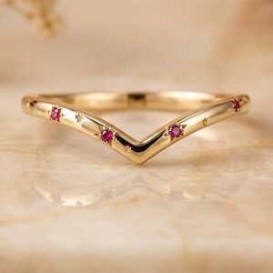 May include: A gold V-shaped chevron ring featuring three small round-cut pink rubies set into the band. The metal surface is polished and includes engraved star-shaped details surrounding each of the three inset gemstones.