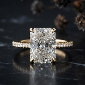 Op de afbeelding: Een ring met een radiant-geslepen diamant in een geelgouden band. De centrale rechthoekige steen wordt vastgehouden door vier klauwen en de band is voorzien van een pavé-zetting met kleine ronde diamanten langs de zijkanten.