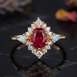 May include: A gold ring featuring a central oval-cut deep red gemstone surrounded by a halo of small round white stones. Two pear-shaped iridescent opals are set on the sides of the band, accented by small square-cut stones at the top and bottom.