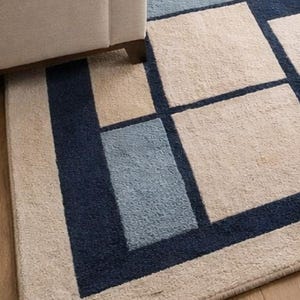 May include: Geometric patterned rug with a beige, navy blue, light blue, and yellow design. The rug is rectangular and features a repeating pattern of squares and rectangles. The rug is placed on a wooden floor, with a beige sofa in the background.