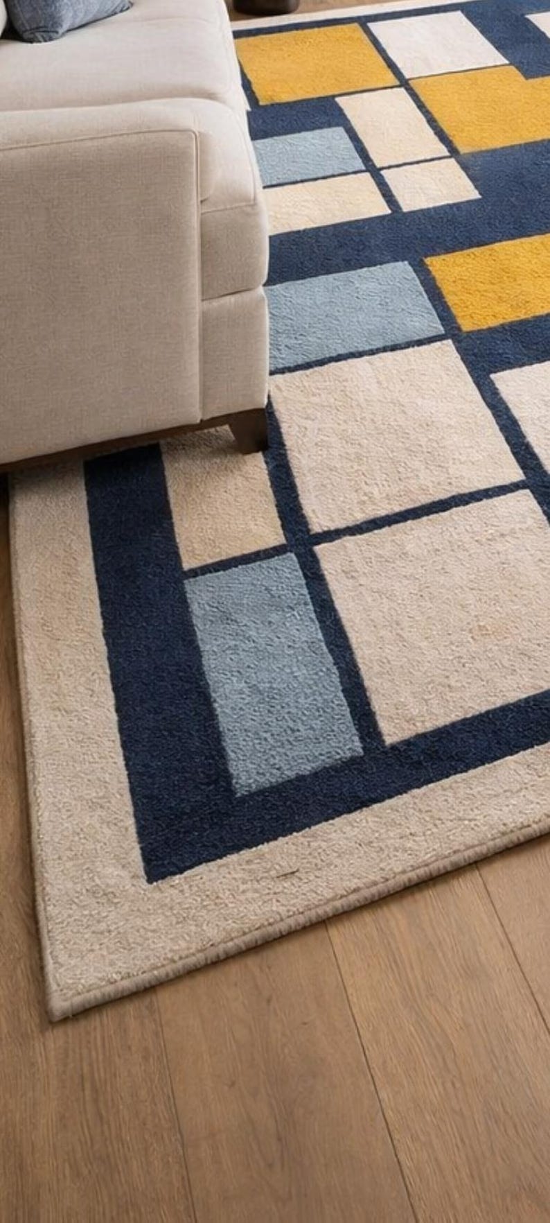 May include: Geometric patterned rug with a beige, navy blue, light blue, and yellow design. The rug is rectangular and features a repeating pattern of squares and rectangles. The rug is placed on a wooden floor, with a beige sofa in the background.