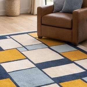 May include: Geometric patterned area rug with a modern design. The rug features a grid of squares in navy blue, mustard yellow, light blue, and cream colors. The rug is placed in a living room setting with a brown leather armchair.