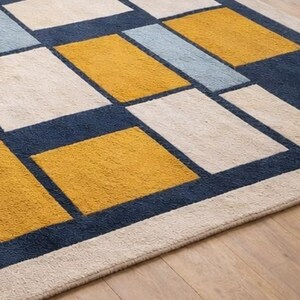May include: A rectangular area rug with a geometric pattern. The rug features squares in cream, navy blue, light blue, and mustard yellow. The rug is placed on a hardwood floor.