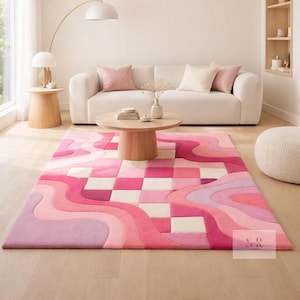 Op de afbeelding: Een roze en witte vloerkleed met een golvend patroon en dambord secties. Het vloerkleed ligt in een woonkamer met een witte bank, een houten salontafel en een vloerlamp.