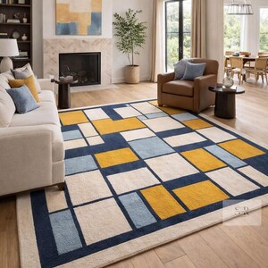May include: A modern area rug with a geometric design. The rug features a pattern of rectangles in navy blue, cream, light blue, and yellow. The rug is placed in a living room with a white sofa and brown leather armchair.