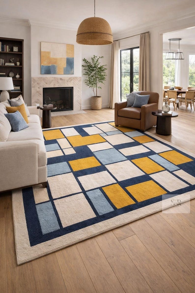 May include: A modern area rug with a geometric design. The rug features a pattern of rectangles in navy blue, cream, light blue, and yellow. The rug is placed in a living room with a white sofa and brown leather armchair.