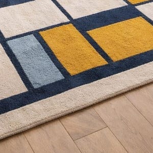 May include: Geometric patterned rug featuring a grid of rectangles in navy blue, cream, light blue, and mustard yellow. The rug is placed on a hardwood floor.