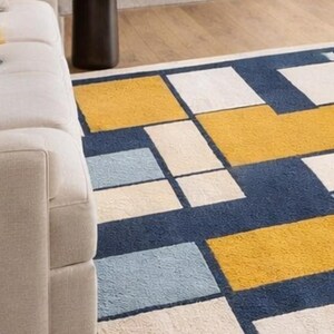 May include: A geometric patterned rug with a modern design. The rug features large rectangular blocks in navy blue, yellow, light blue, and cream colors. The rug is placed in a living room setting, with a white sofa visible in the frame.