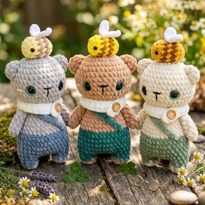 Puede incluir: Tres osos de peluche de ganchillo hechos a mano con sombreros de abeja. Los osos son de color gris, marrón y crema, con trajes azules, verdes y crema. Tienen detalles de botones y están colocados sobre una superficie de madera con flores.