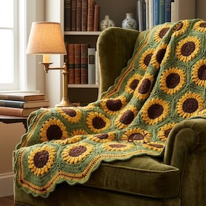May include: A crochet blanket featuring a repeating pattern of sunflower motifs with dark brown centres and bright yellow petals, connected by a sage green background. The blanket is draped over a green upholstered armchair, with a pair of glasses.