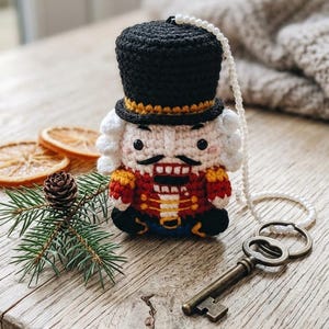 Könnte beinhalten: Gehäkelte Nussknacker-Puppe mit schwarzem Zylinder, roter Jacke und weißem Haar. Die Puppe hat eine Perlenkette und sitzt auf einer Holzoberfläche mit einem antiken Schlüssel und einem Tannenzweig. Eine festliche Weihnachtsdekoration.