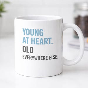 May include: White ceramic mug with the text "YOUNG AT HEART. OLD EVERYWHERE ELSE." in blue and black. The mug has a curved handle and is set on a white surface. A small plant and cookies are in the background.