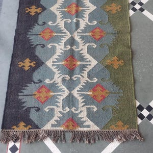 May include: Rectangular woven rug with a geometric pattern in shades of blue, green, and beige. The rug features a central design with diamond and zigzag shapes, accented with red and gold details. It has a fringed edge.