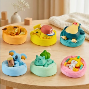 May include: A collection of six colorful figurines of popular characters, each resting on a small, round cushion. The figurines include a yellow Pikachu wearing a baseball cap, a blue Snorlax, and a sleeping Eevee. The cushions are in various colors.
