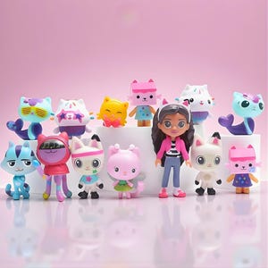 Può includere: Una collezione di colorate statuette di Gabby's Dollhouse, tra cui personaggi a tema gatto e una bambola. Le figure sono in varie pose e design, con uno sfondo rosa. La bambola indossa una giacca rosa e jeans blu.